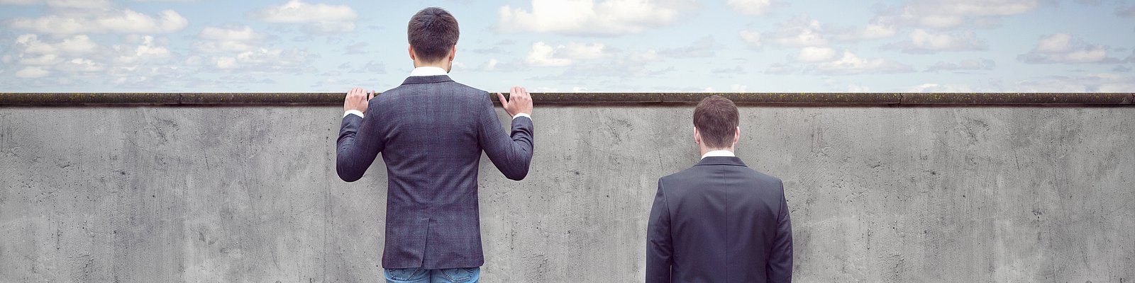 Zwei Männer stehen vor einer Mauer, einer davon auf einem Bücherstapel schaut über die Mauer hinweg