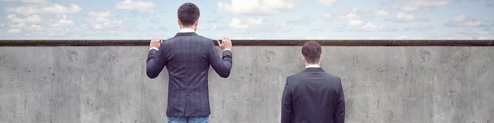 Zwei Männer stehen vor einer Mauer, einer davon auf einem Bücherstapel schaut über die Mauer hinweg